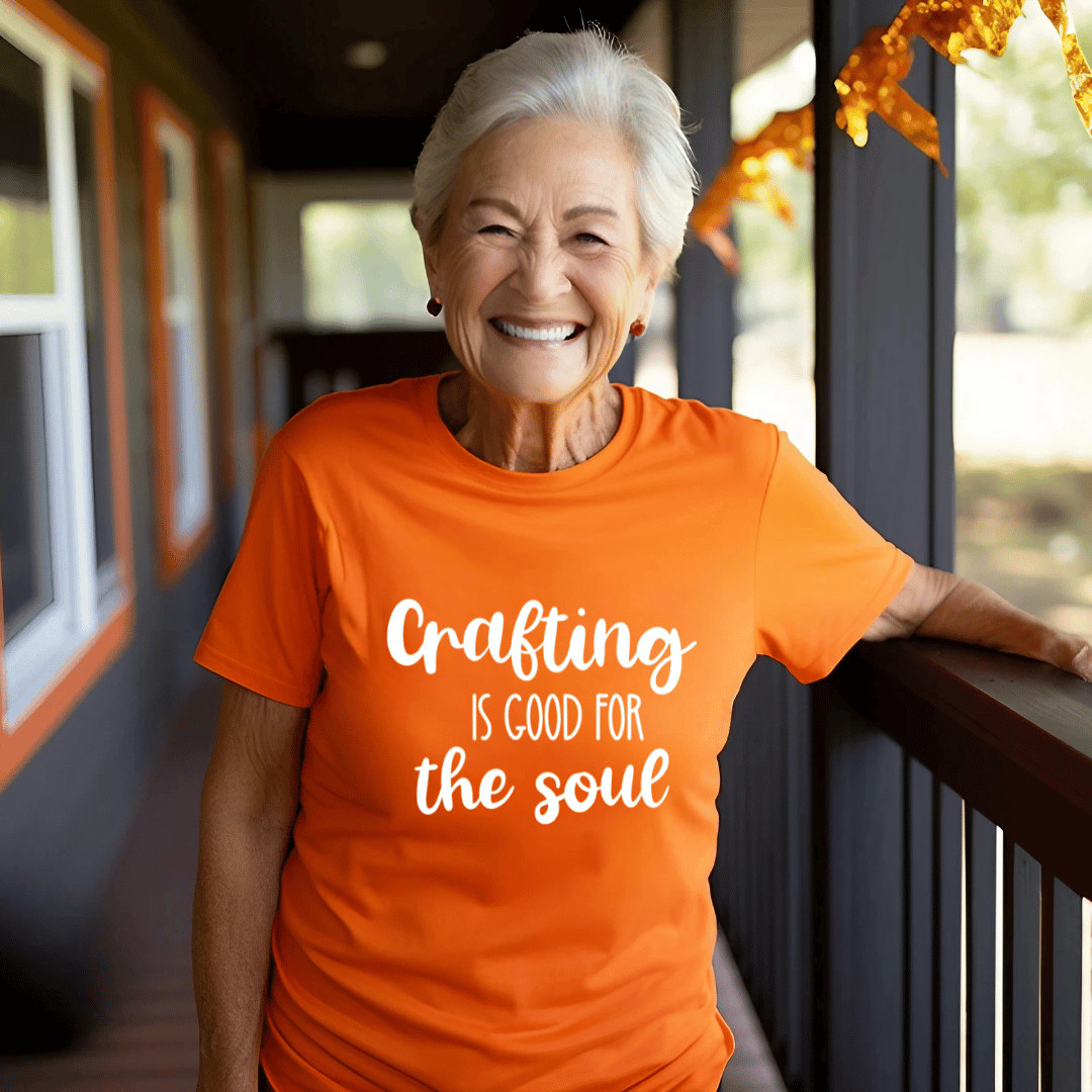 Woman wearing an orange t-shirt with 'Crafting is good for the soul' text, standing on a porch.