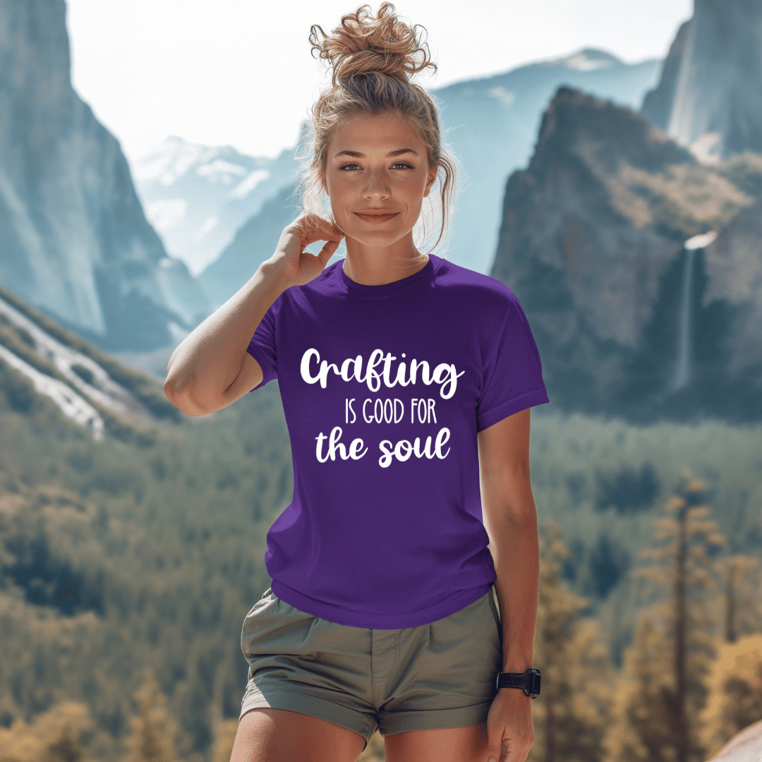 Woman wearing a purple t-shirt with 'Crafting is good for the soul' text, standing in front of mountains.