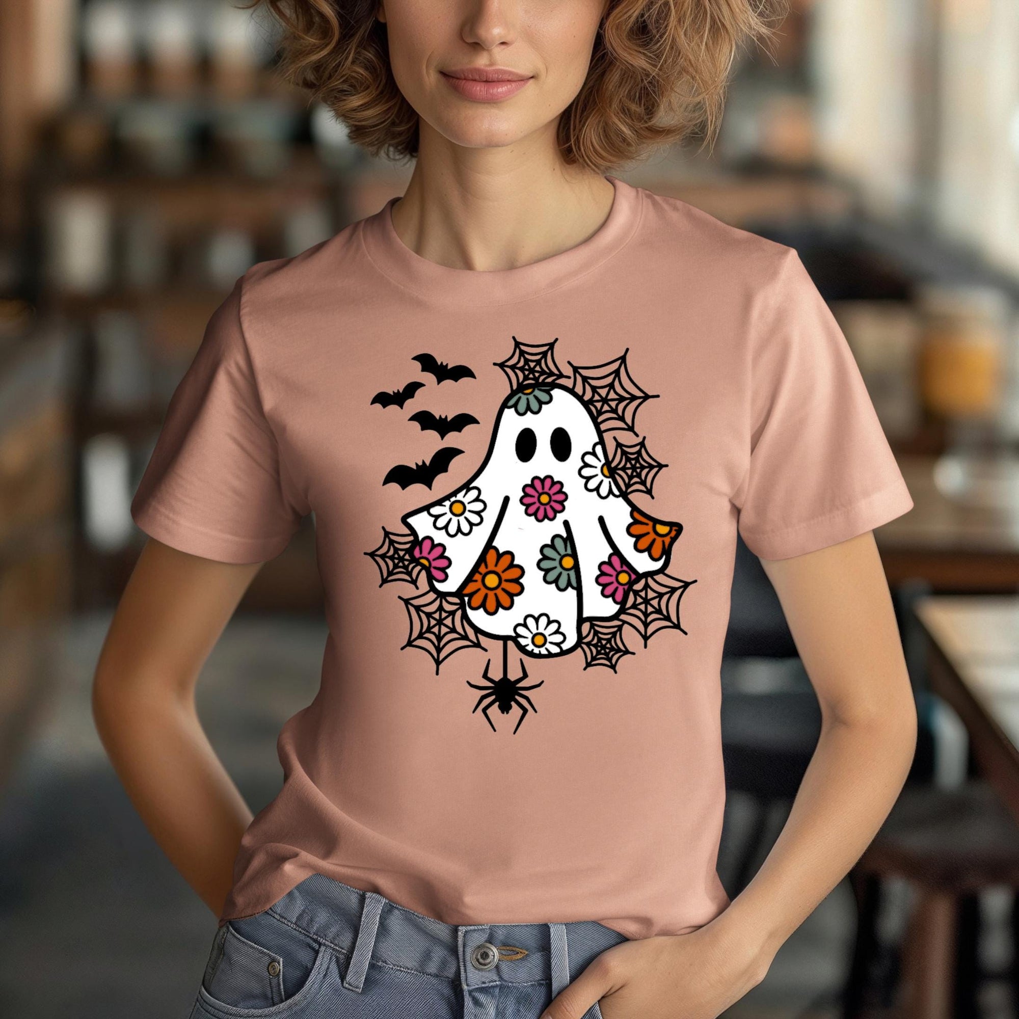 A woman wearing a peach t-shirt with a whimsical design featuring a ghost, bats, and spiders on it.