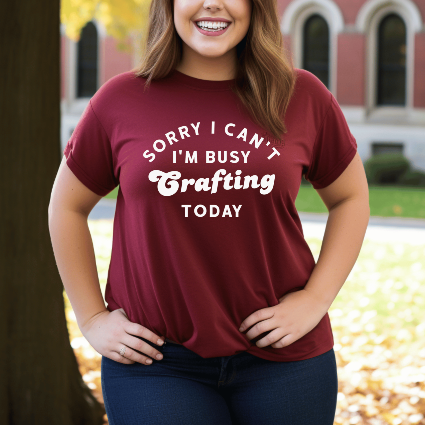 Woman wearing a maroon t-shirt with text in front of a building with autumn foliage.