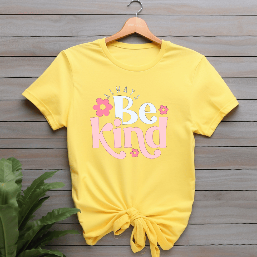 Yellow t-shirt with 'Always Be Kind' text on a wooden surface with a plant.