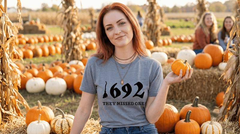Woman holding a gourd while sitting on a hay bale in a pumpkin farm wearing a tshirt that says 1692 They Missed One