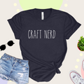 A black t-shirt with 'CRAFT NERD' text in white on the front, displayed with denim shorts, sunglasses, and a potted plant in the background.