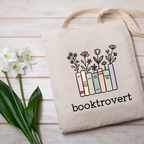 Tote bag with 'booktrovert' design featuring books and flowers on a wooden surface with flowers.