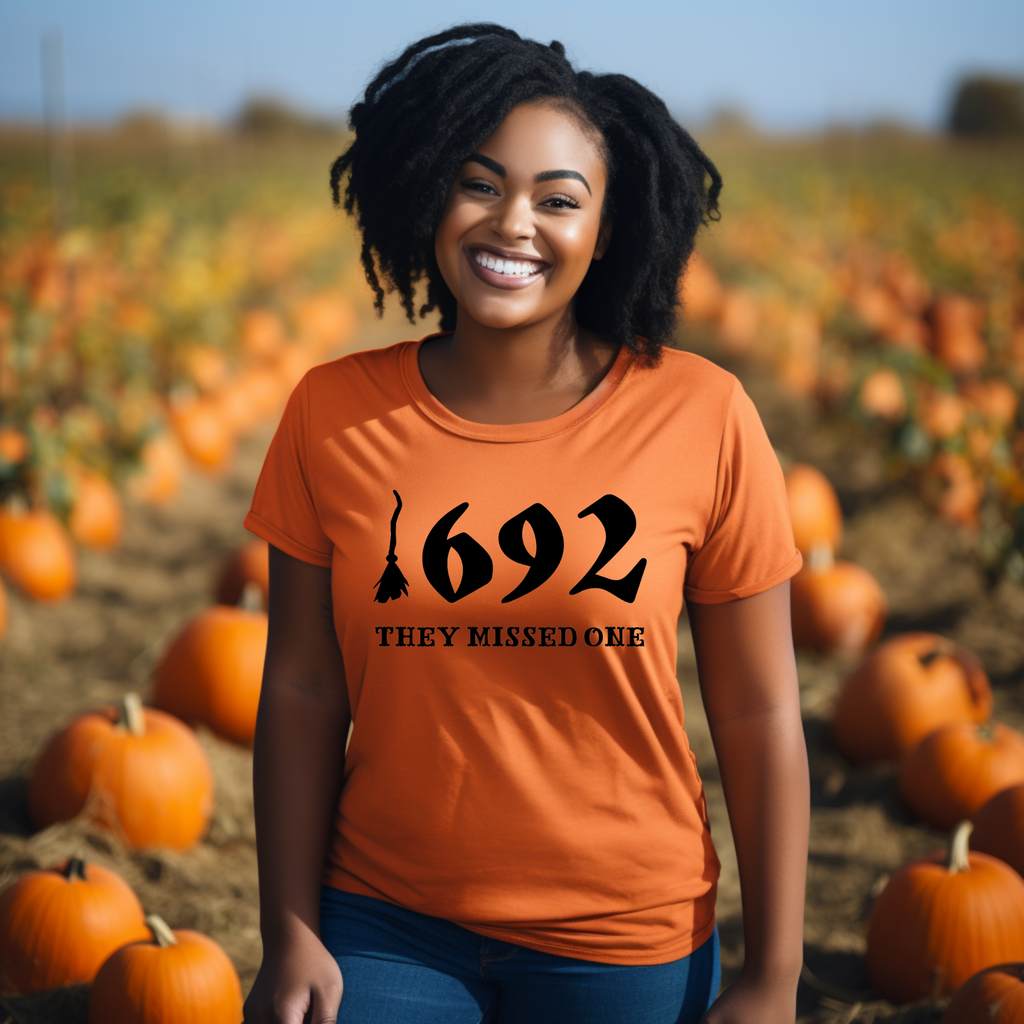 A person in a pumpkin patch wearing an orange t-shirt with the text '1692 THEY MISSED ONE' on it.