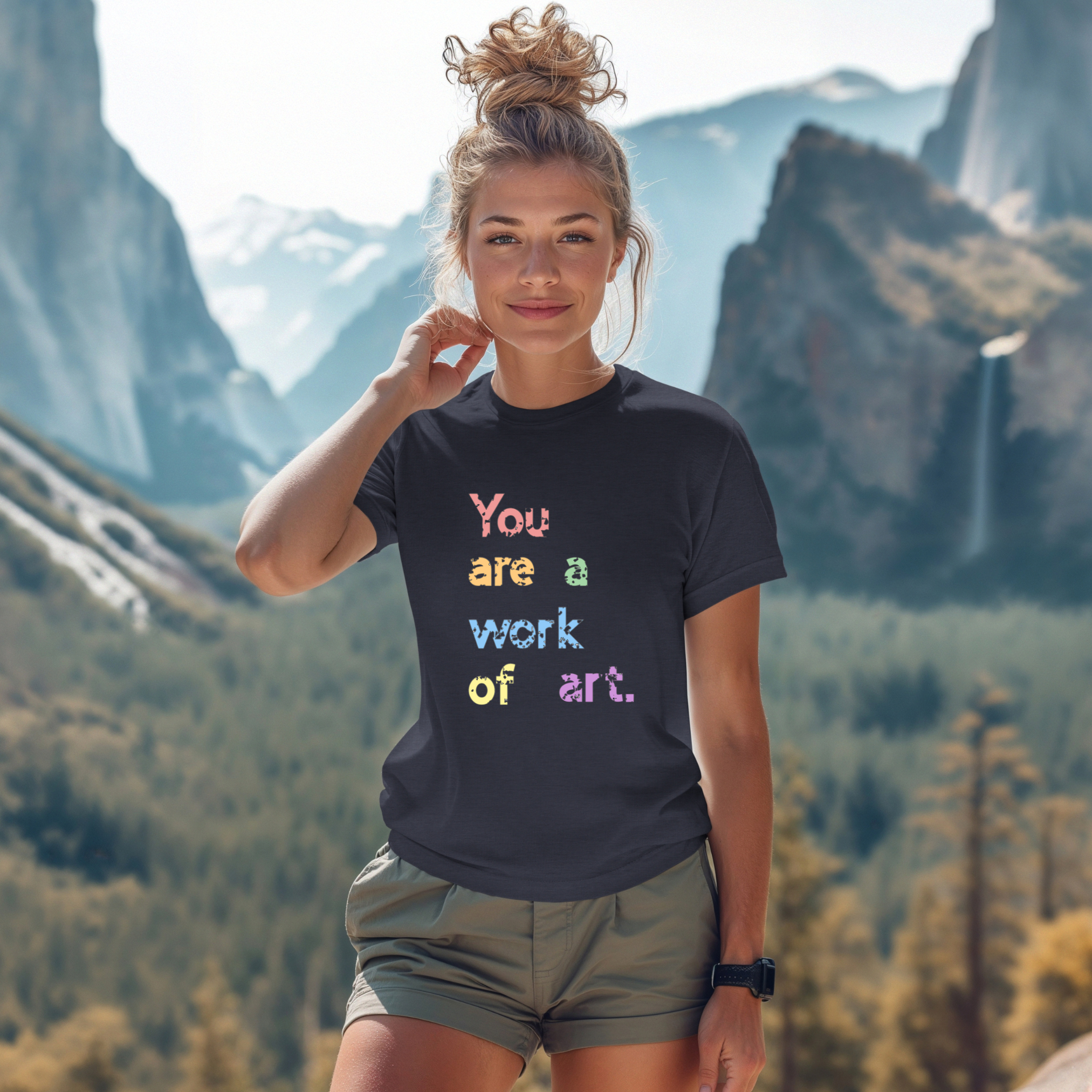 A person wearing a dark grey t-shirt with the text 'You are a work of art' printed in colorful letters, standing in a scenic mountainous landscape.