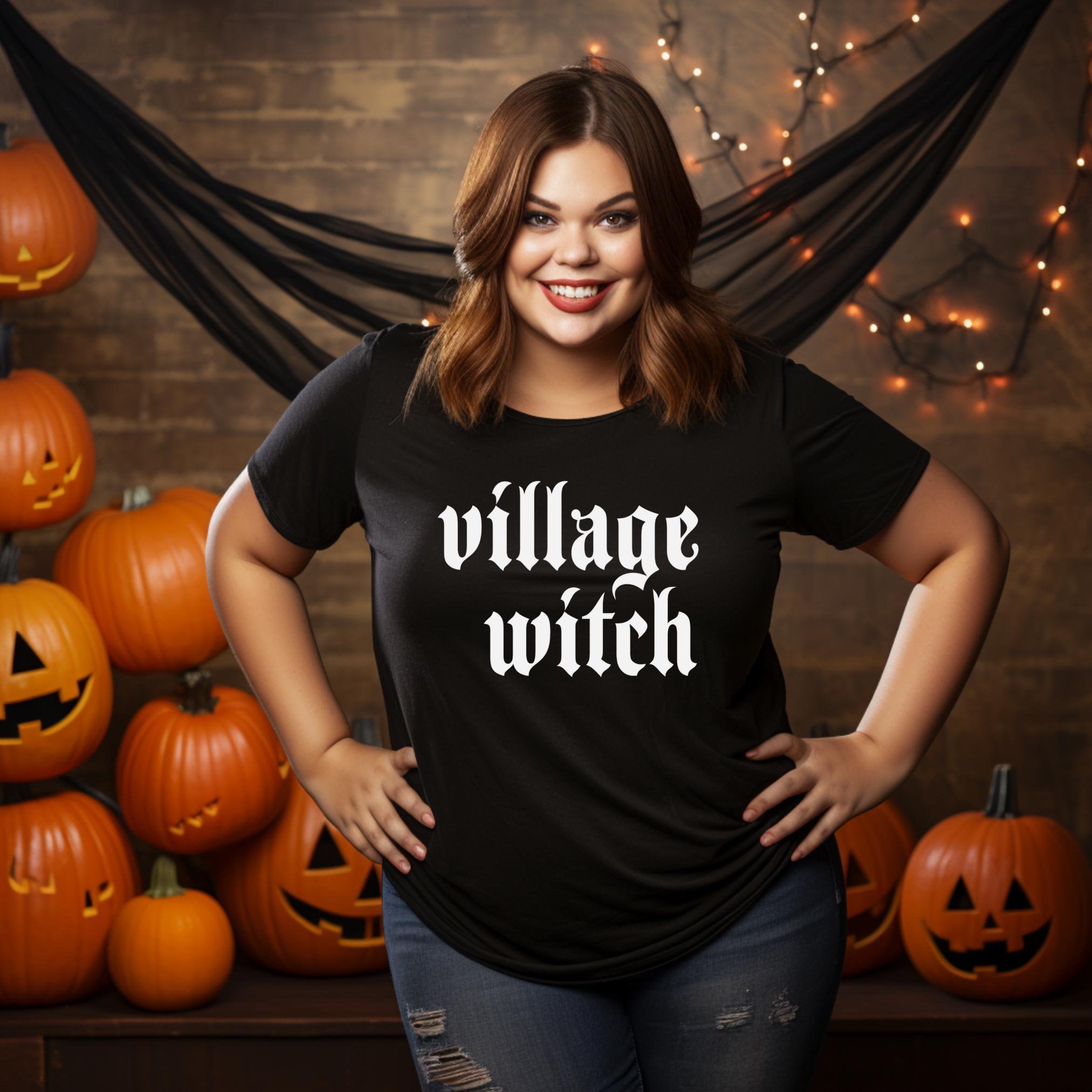 A woman standing in front of a backdrop with pumpkins and a text that reads 'VILLAGE WITCH'. She is wearing a black t-shirt with the same text.