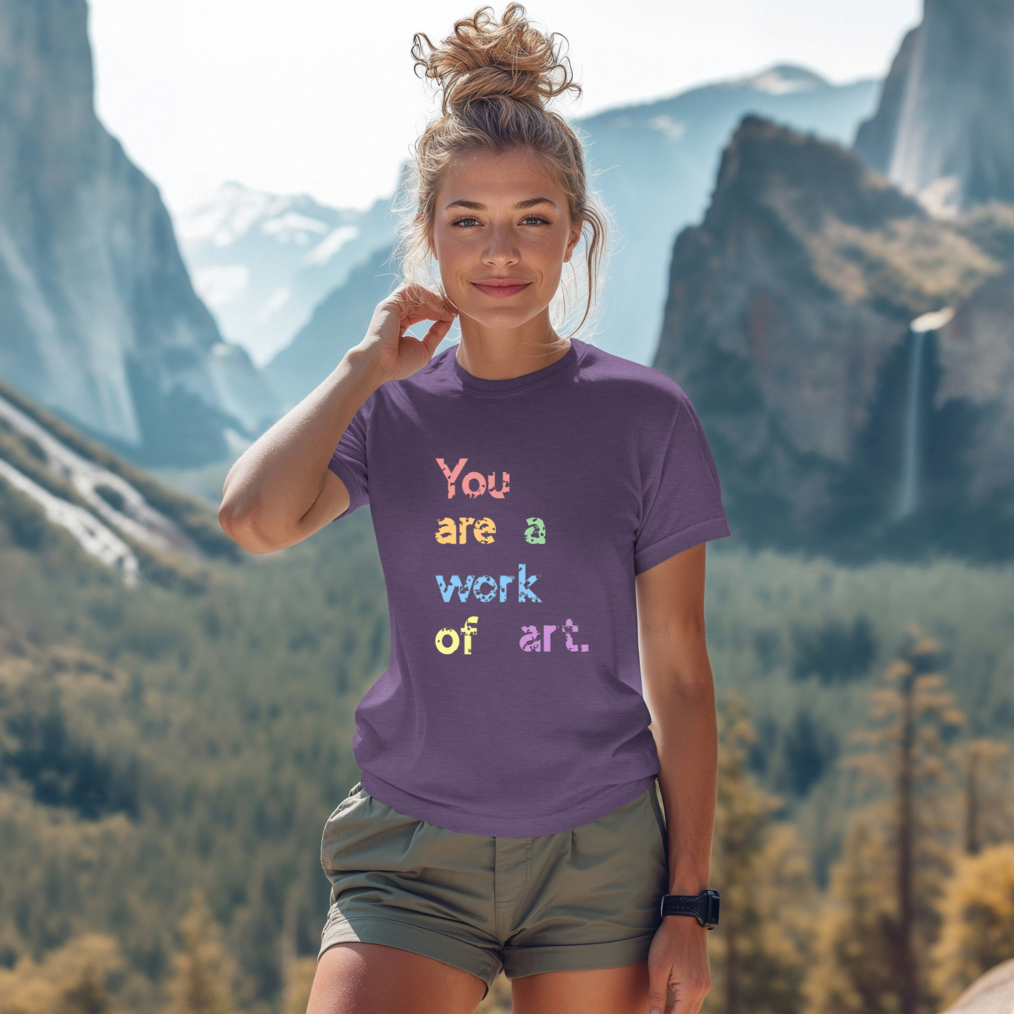 A person wearing a purple t-shirt with the text 'You are a work of art' printed in colorful letters, standing in a scenic mountainous landscape.