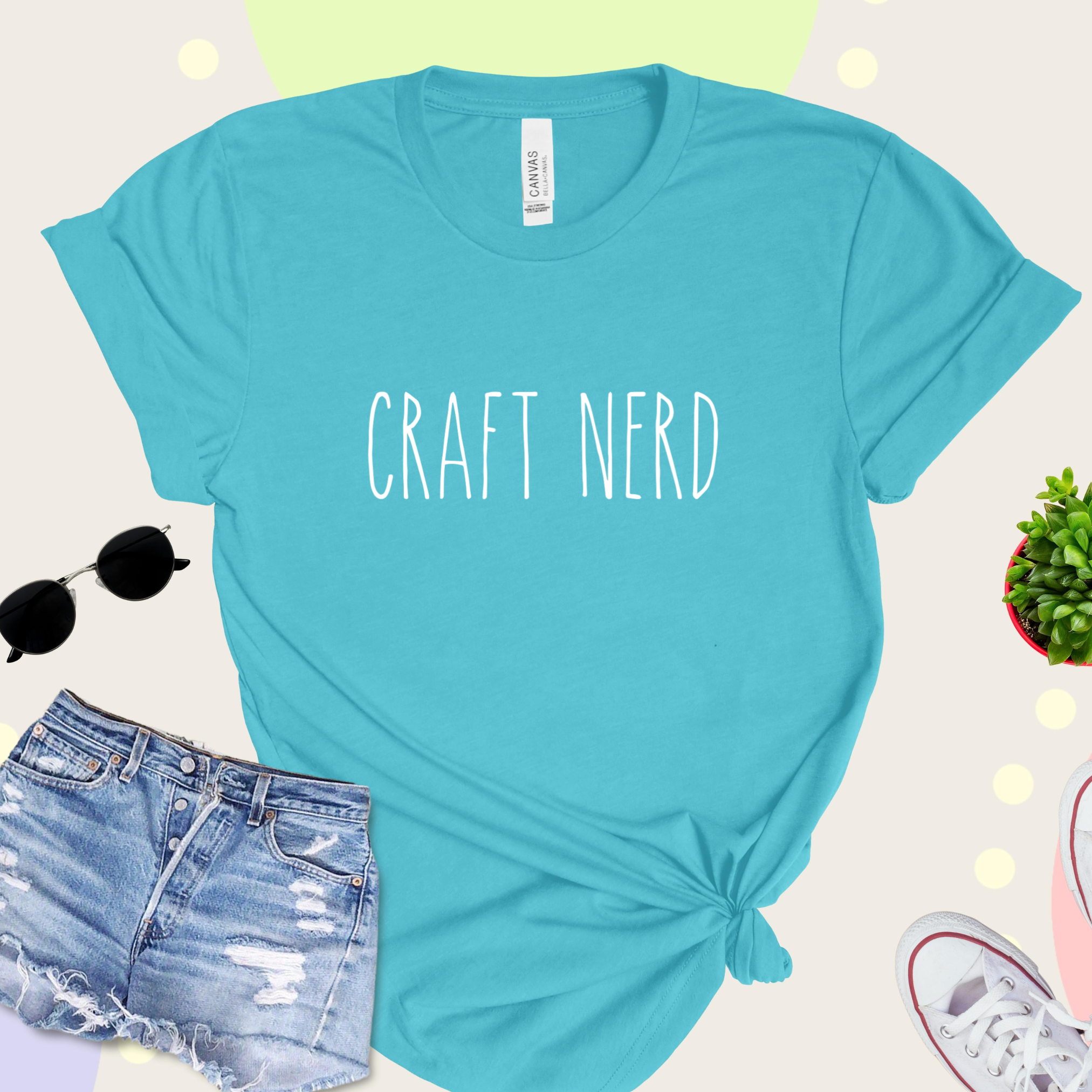 A teal t-shirt with 'CRAFT NERD' text in white on the front, displayed with denim shorts, sunglasses, and a potted plant in the background.