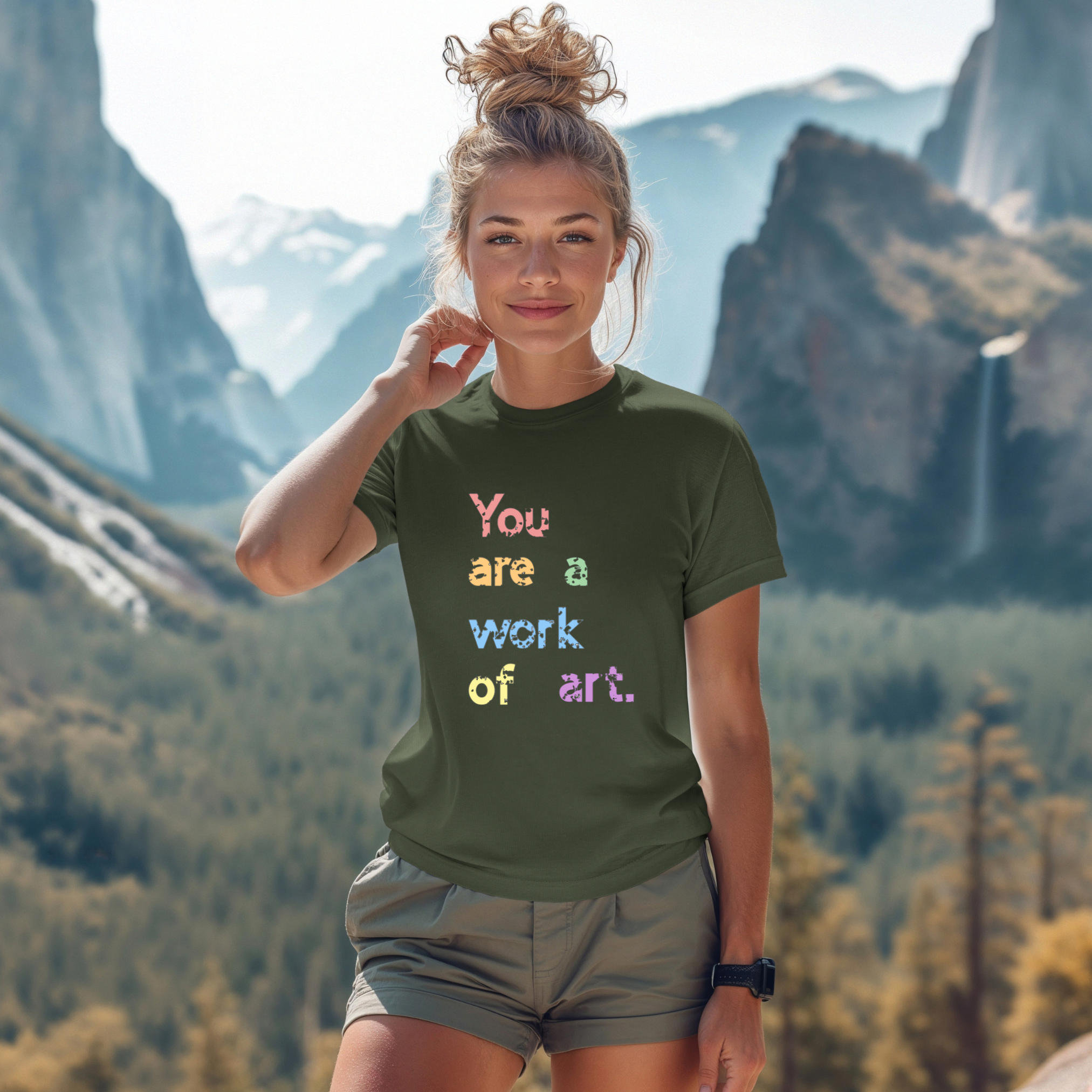 A person wearing a green t-shirt with the text 'You are a work of art' printed in colorful letters, standing in a scenic mountainous landscape.