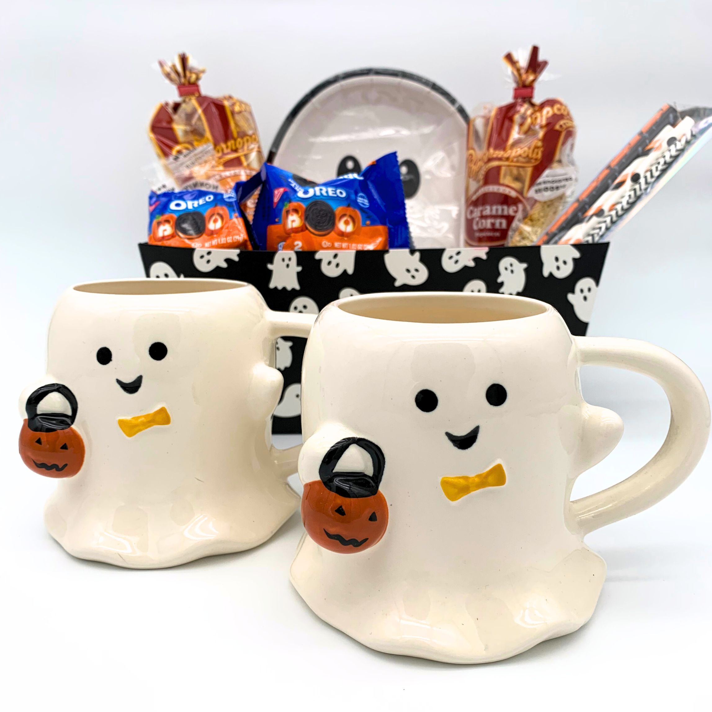 Two ghost-shaped mugs with pumpkin baskets on a white background, with Halloween-themed treats in the background.