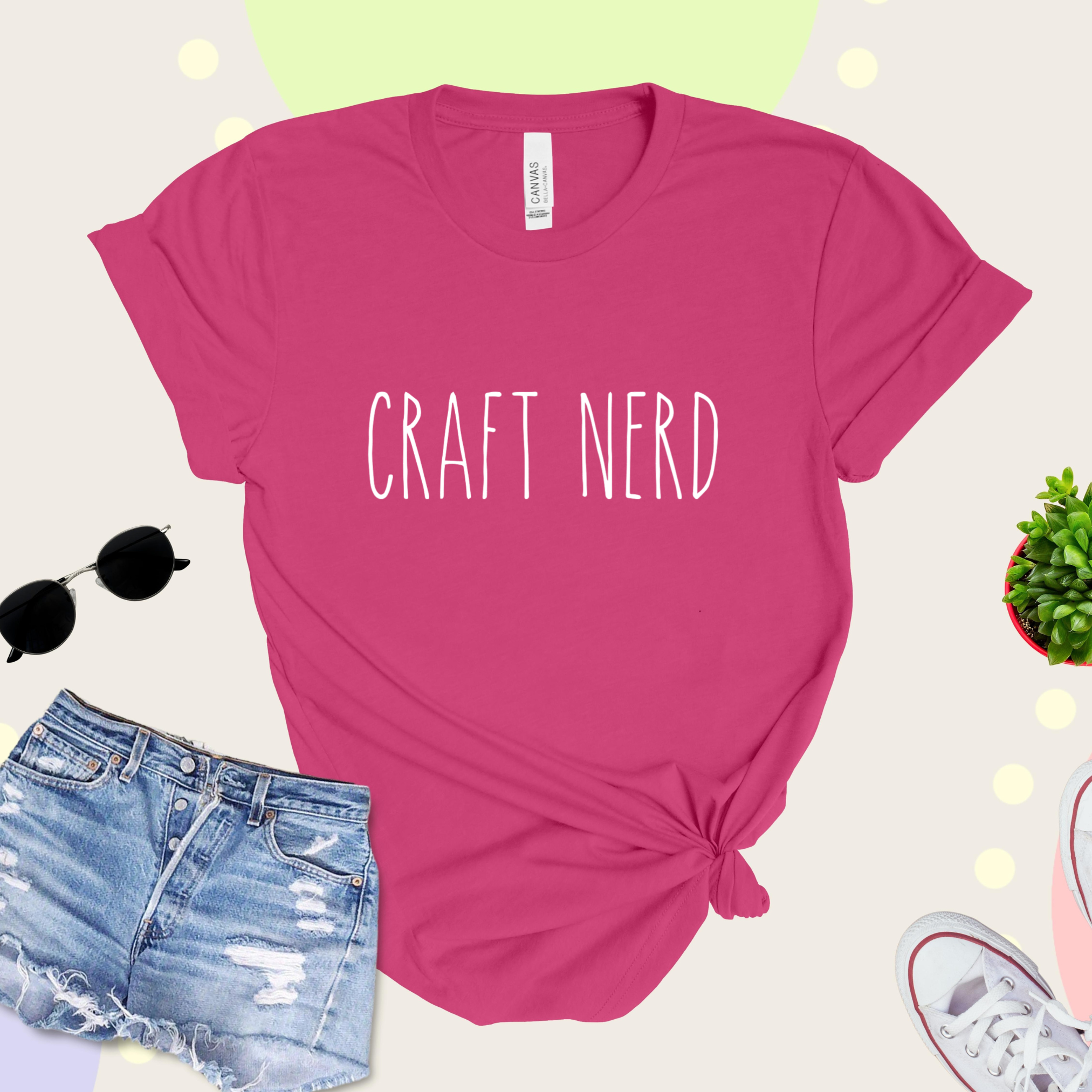A pink t-shirt with 'CRAFT NERD' text in white on the front, displayed with denim shorts, sunglasses, and a potted plant in the background.
