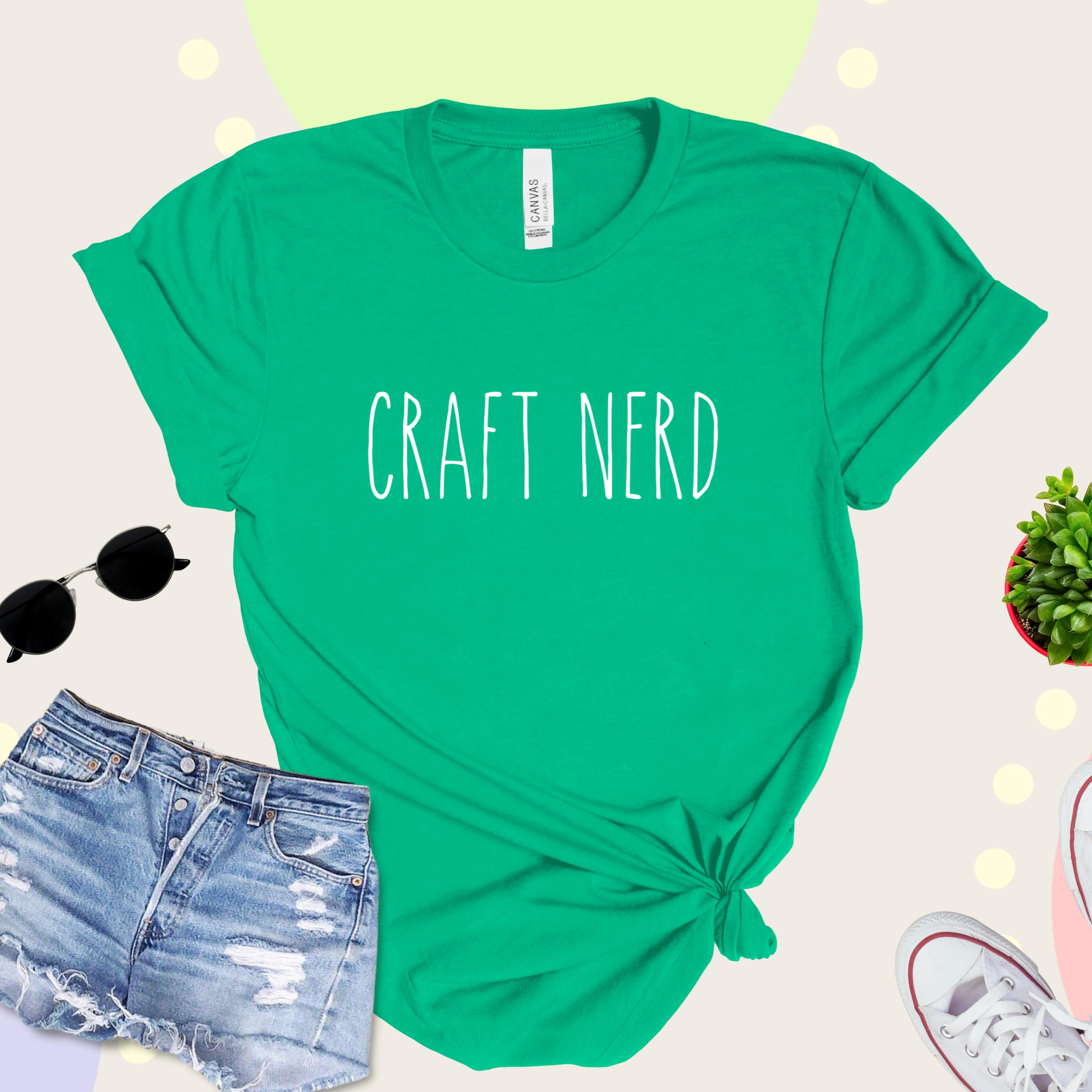 A green t-shirt with 'CRAFT NERD' text in white on the front, displayed with denim shorts, sunglasses, and a potted plant in the background.