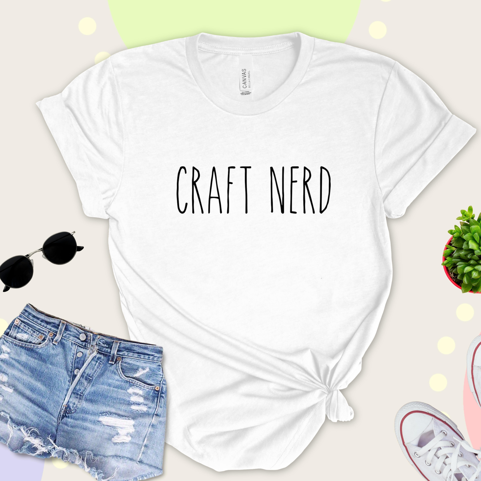 A white t-shirt with 'CRAFT NERD' text in black on the front, displayed with denim shorts, sunglasses, and a potted plant in the background.