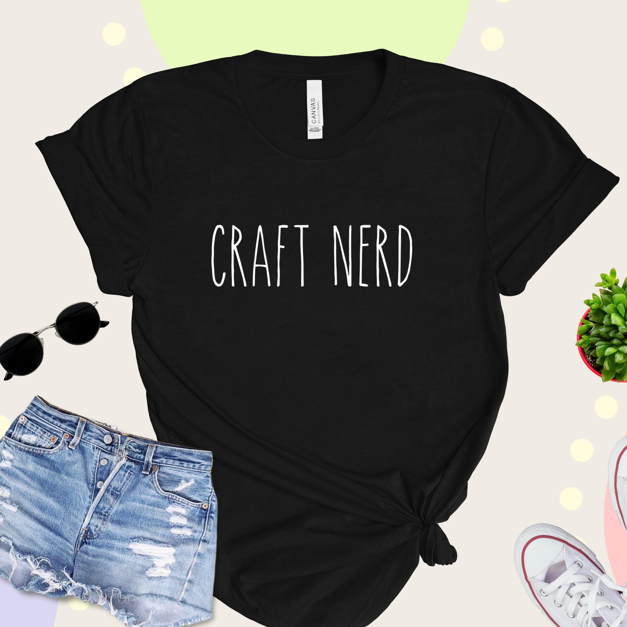 A black t-shirt with 'CRAFT NERD' text in white on the front, displayed with denim shorts, sunglasses, and a potted plant in the background.