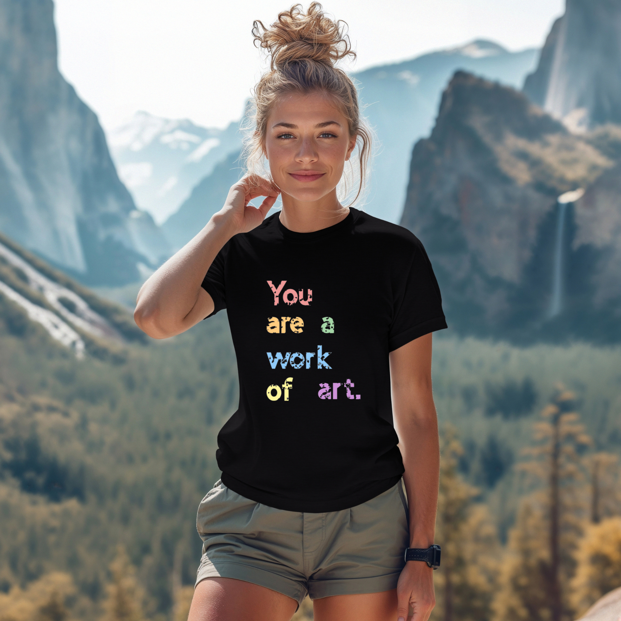A person wearing a black t-shirt with the text 'You are a work of art' printed in colorful letters, standing in a scenic mountainous landscape.