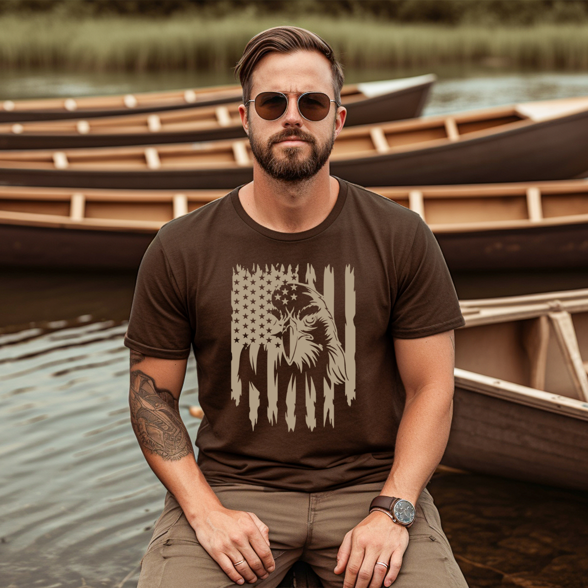 Man wearing a brown t-shirt with a graphic design, sitting by a body of water with canoes in the background.