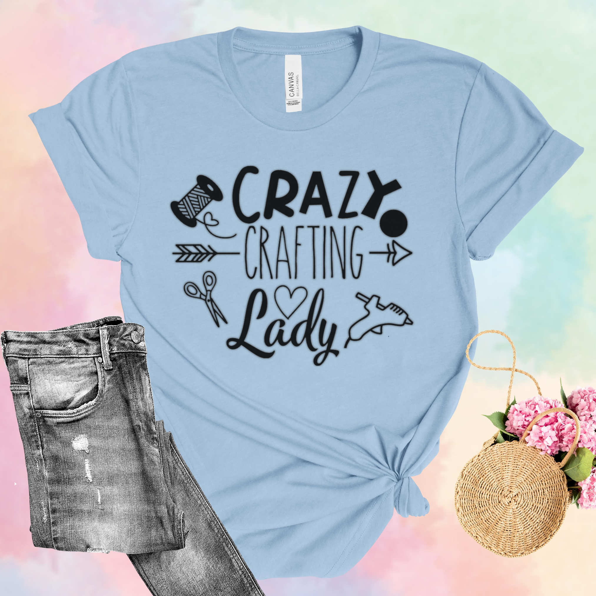 Light blue t-shirt with 'Crazy Crafting Lady' text, paired with jeans and a bag on a colorful background