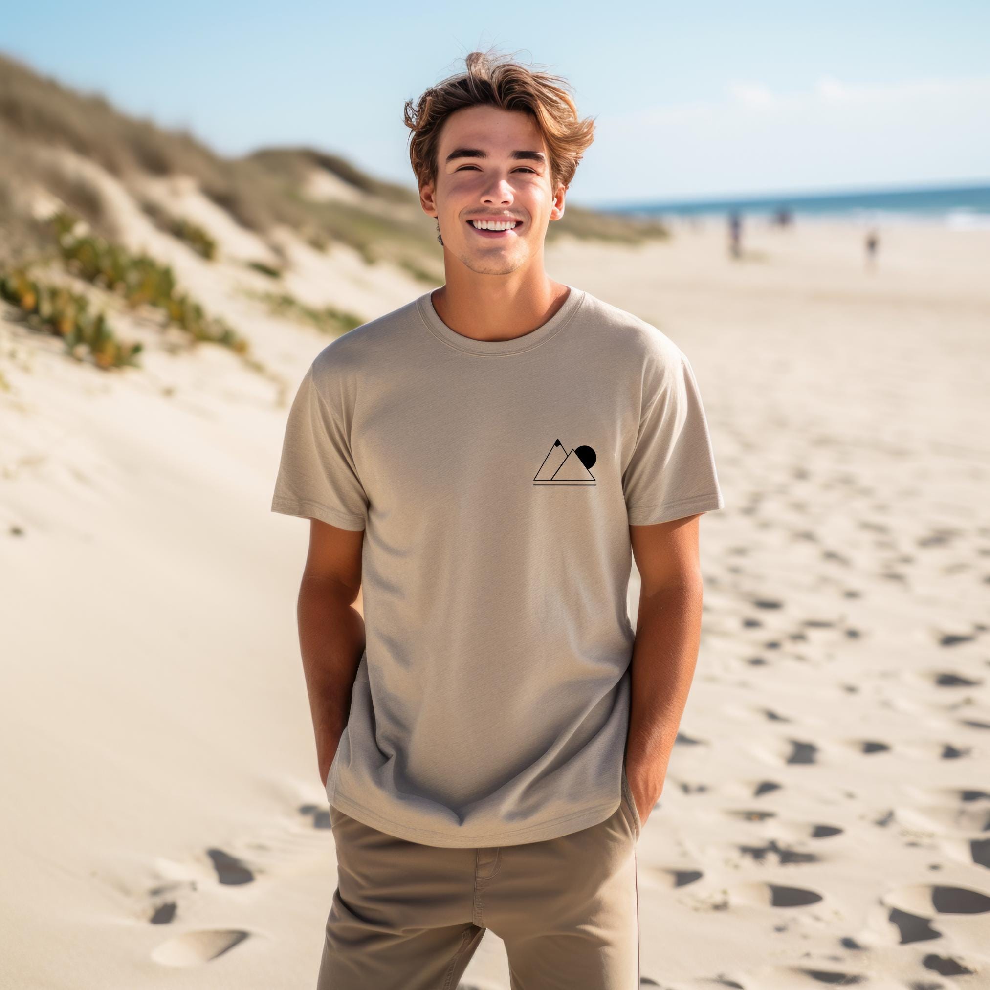A person wearing a tan t-shirt with a simple geometric mountain silhouette on the front, standing on the beach.