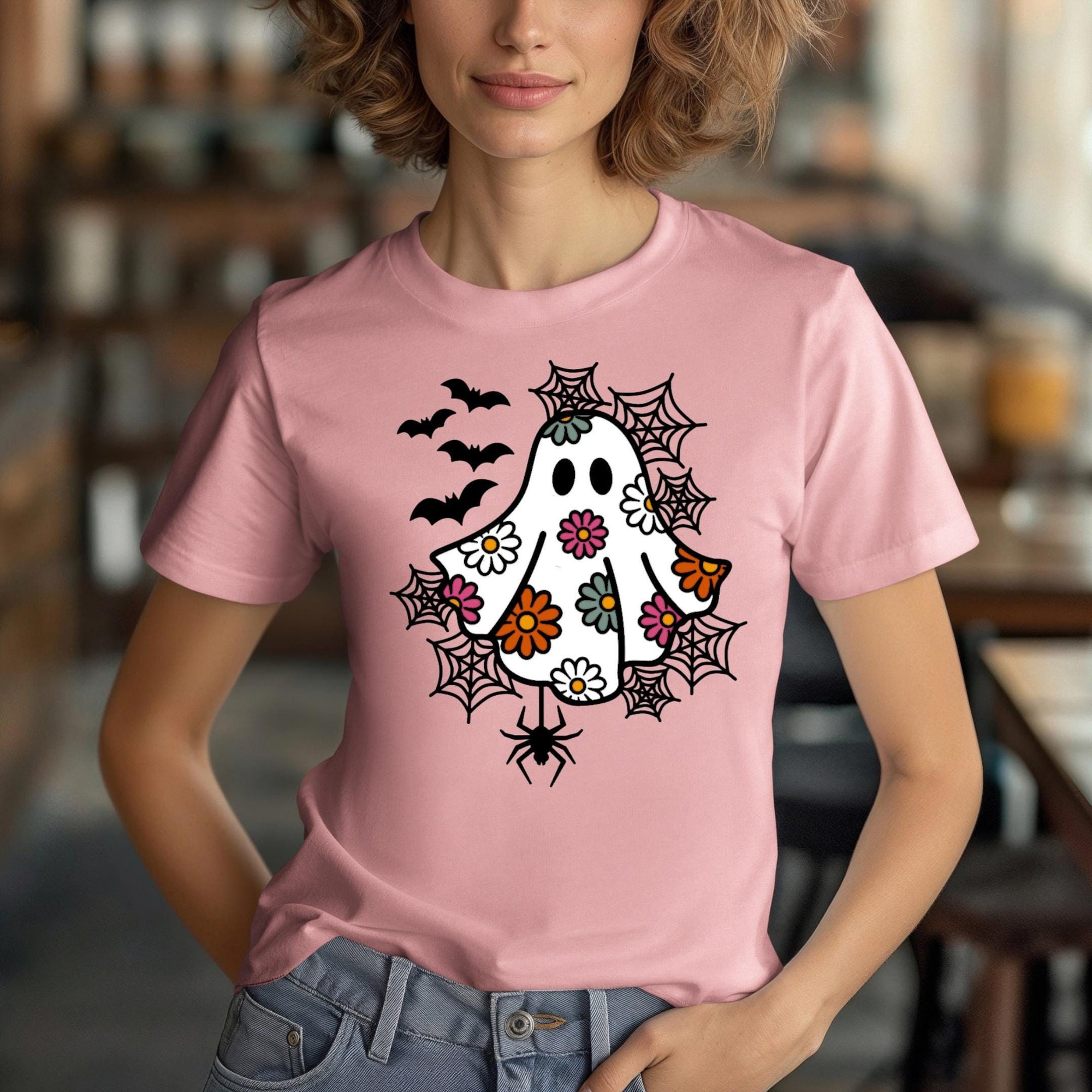 A woman wearing a pink t-shirt with a whimsical design featuring a ghost, bats, and spiders on it.