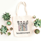Tote bag with 'booktrovert' design surrounded by small potted plants on a white background