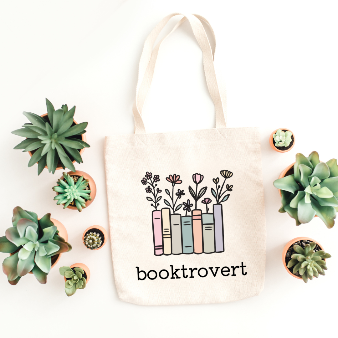 Tote bag with 'booktrovert' design surrounded by small potted plants on a white background