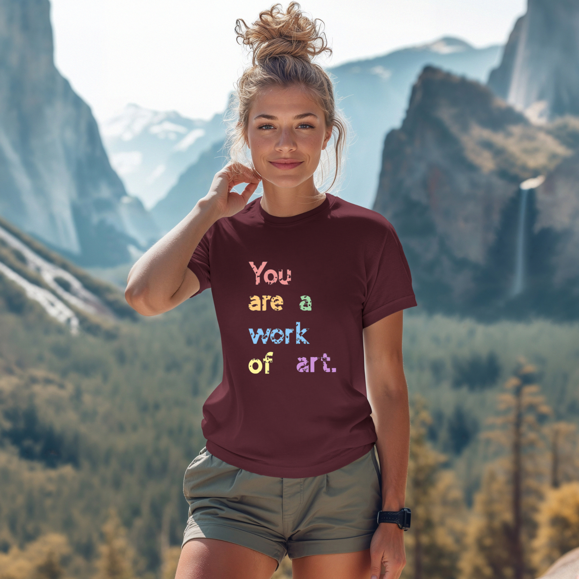 A person wearing a maroon t-shirt with the text 'You are a work of art' printed in colorful letters, standing in a scenic mountainous landscape.