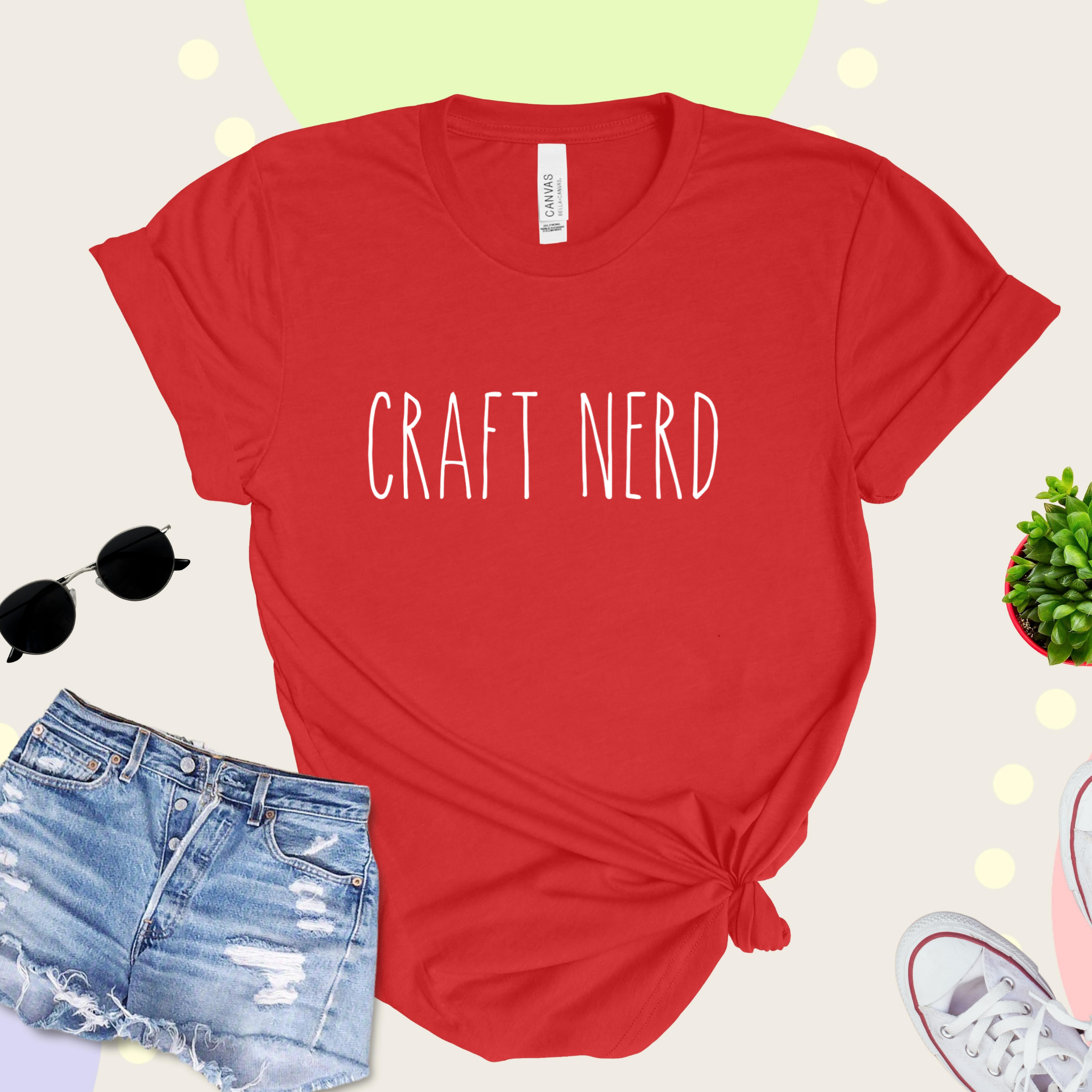 A red t-shirt with 'CRAFT NERD' text in white on the front, displayed with denim shorts, sunglasses, and a potted plant in the background.