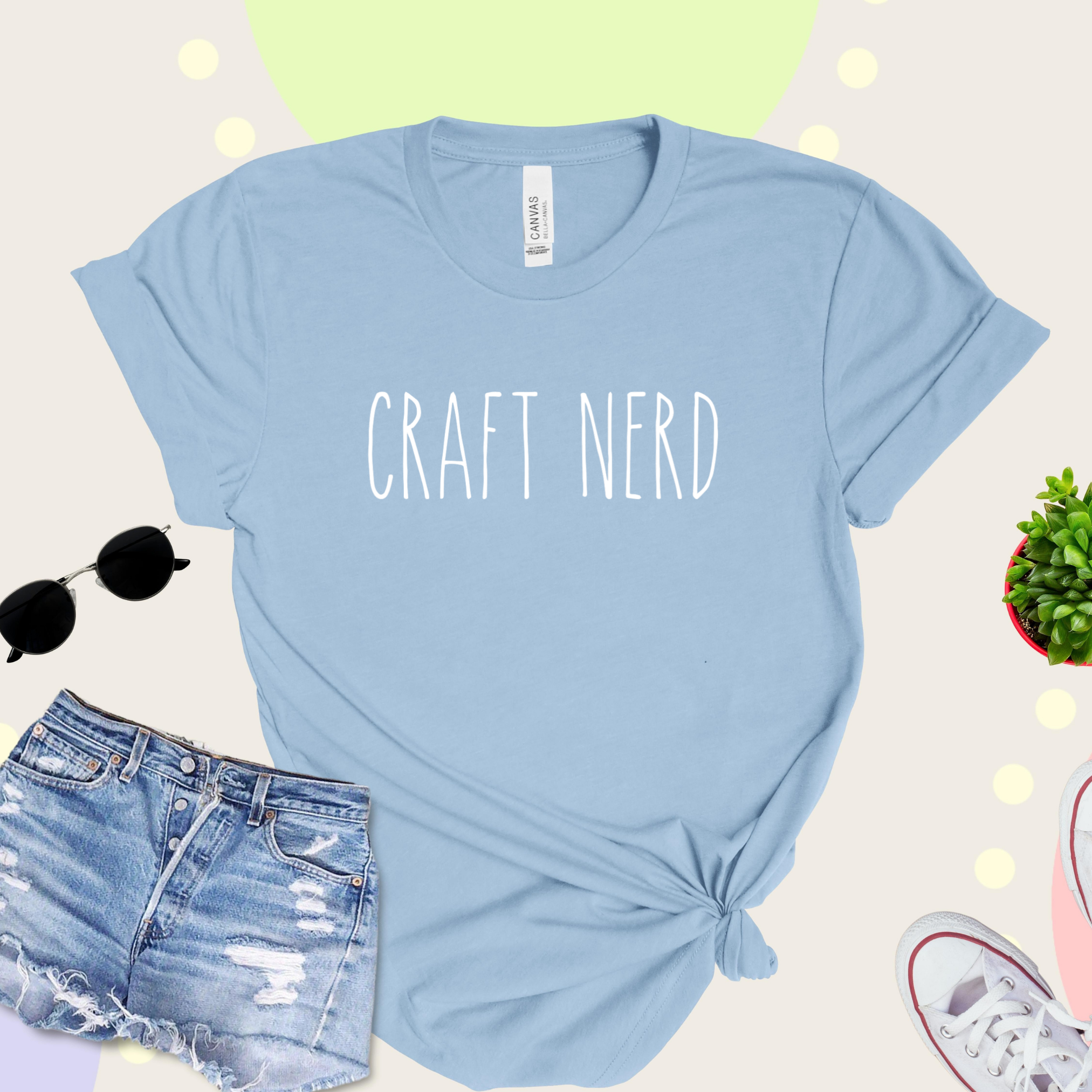 A light blue t-shirt with 'CRAFT NERD' text in white on the front, displayed with denim shorts, sunglasses, and a potted plant in the background.
