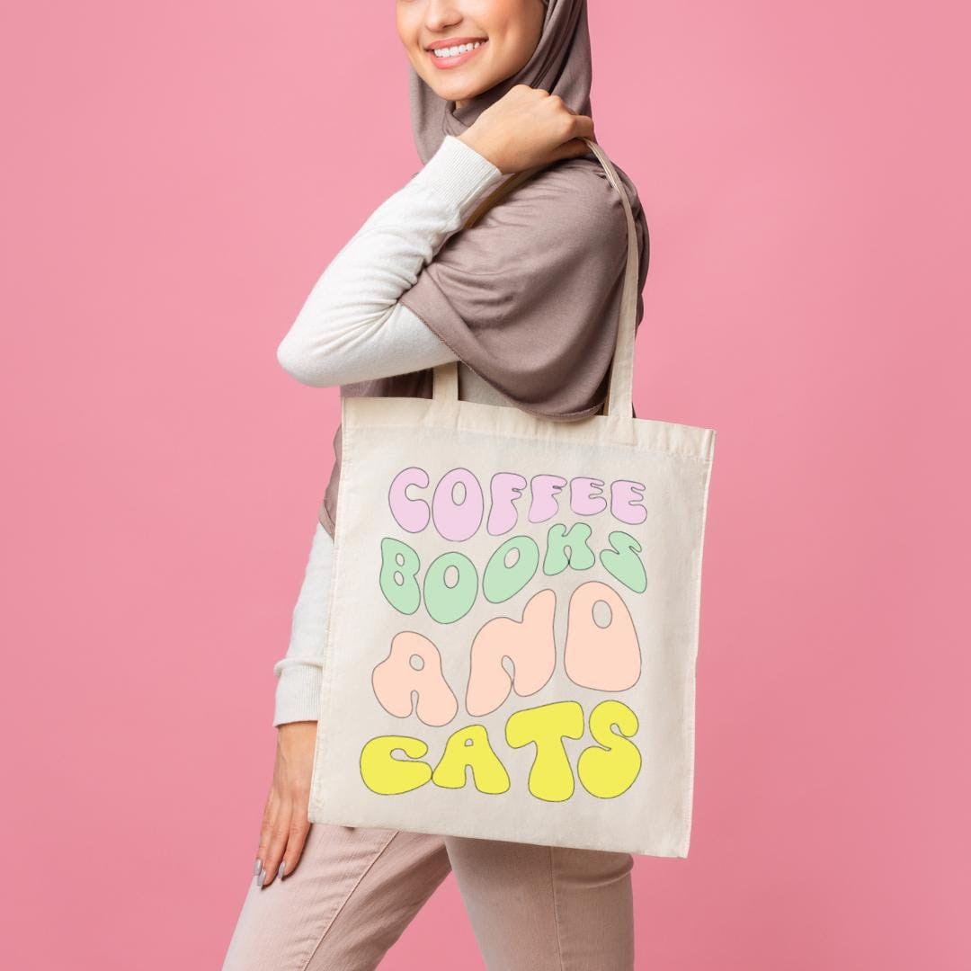 A person holding a beige cotton tote bag with the text 'COFFEE, BOOKS & CATS' printed on the front.