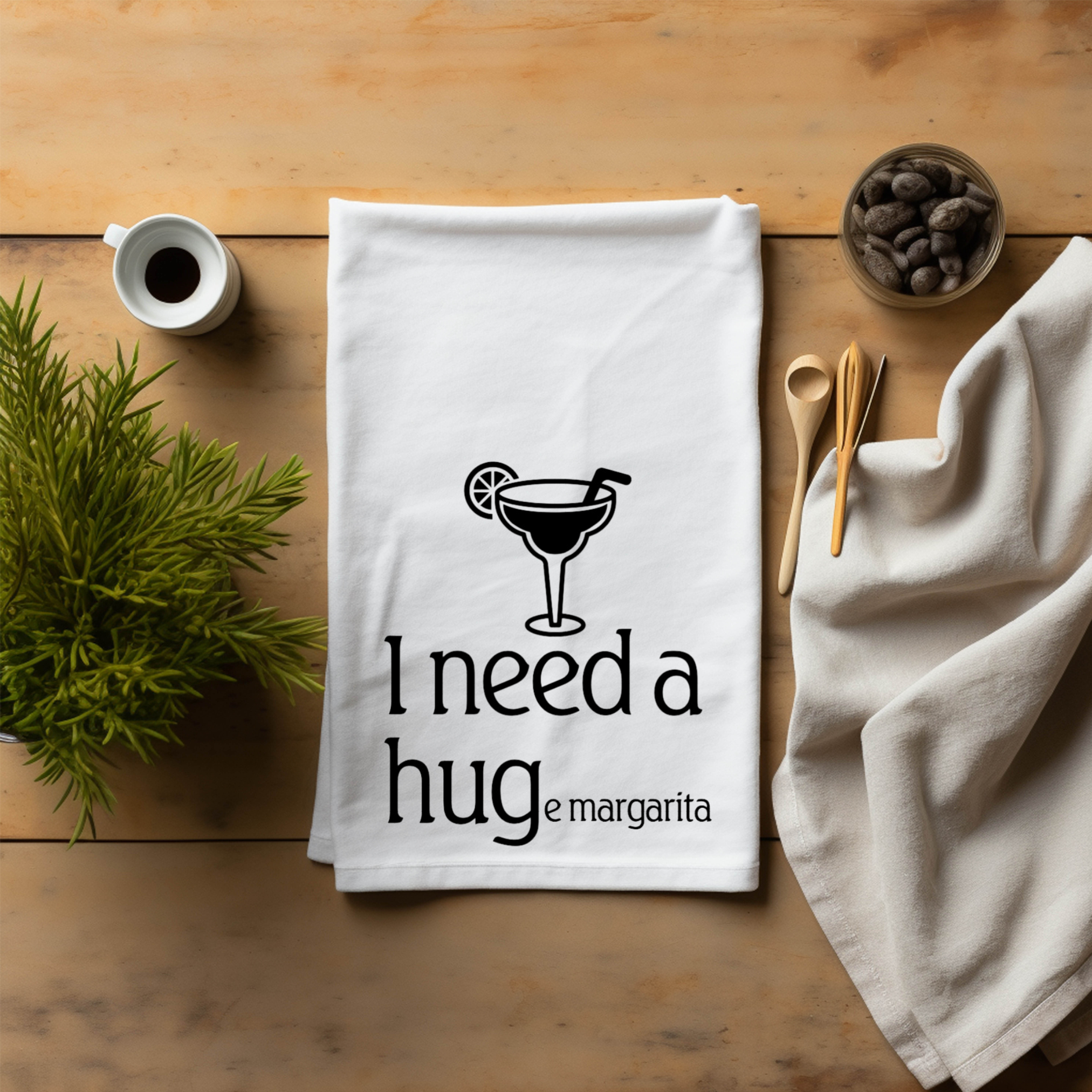 A towel laid flat on a wooden table with kitchen items around it that says 'I need a HUGe margarita'.