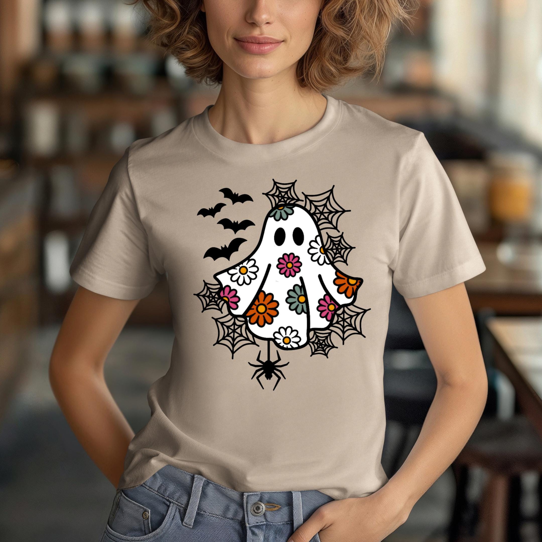 A woman wearing a tan t-shirt with a whimsical design featuring a ghost, bats, and spiders on it.