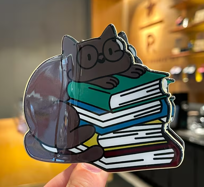 Glossy hair clip of a black cat with glasses on books held in front of a blurred indoor background
