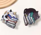 Two cartoon-style hair clips featuring cats in glasses hugging books on a white background