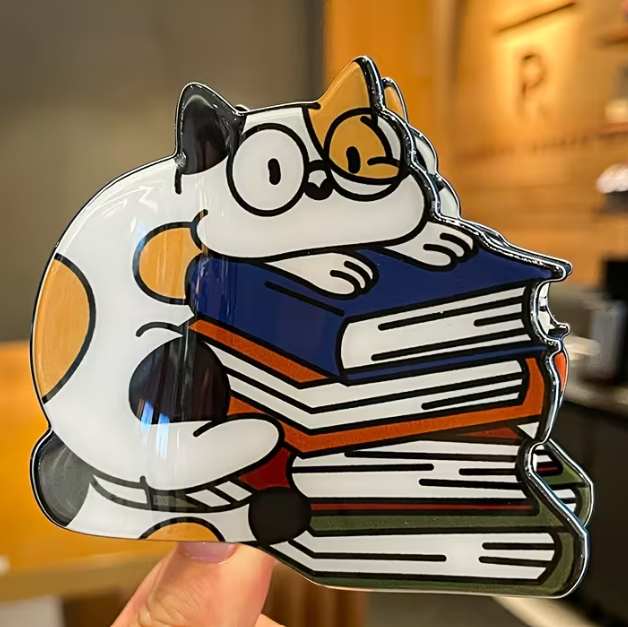 Hair clip featuring a cartoon calico cat holding books with a blurred indoor background