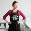 Woman wearing a black apron with a bouquet design features dryer, shears, straightener, spray bottle & comb,