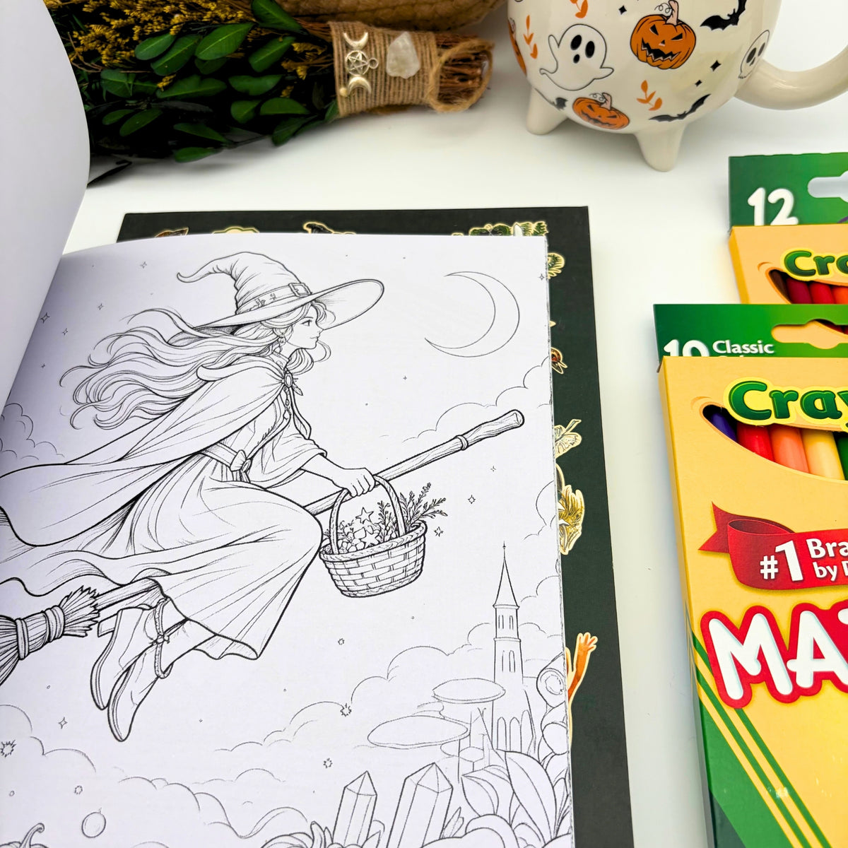Witch coloring book open to a line drawing of a witch flying on a broom past a crescent moon, castle tower, crystals, and mushrooms