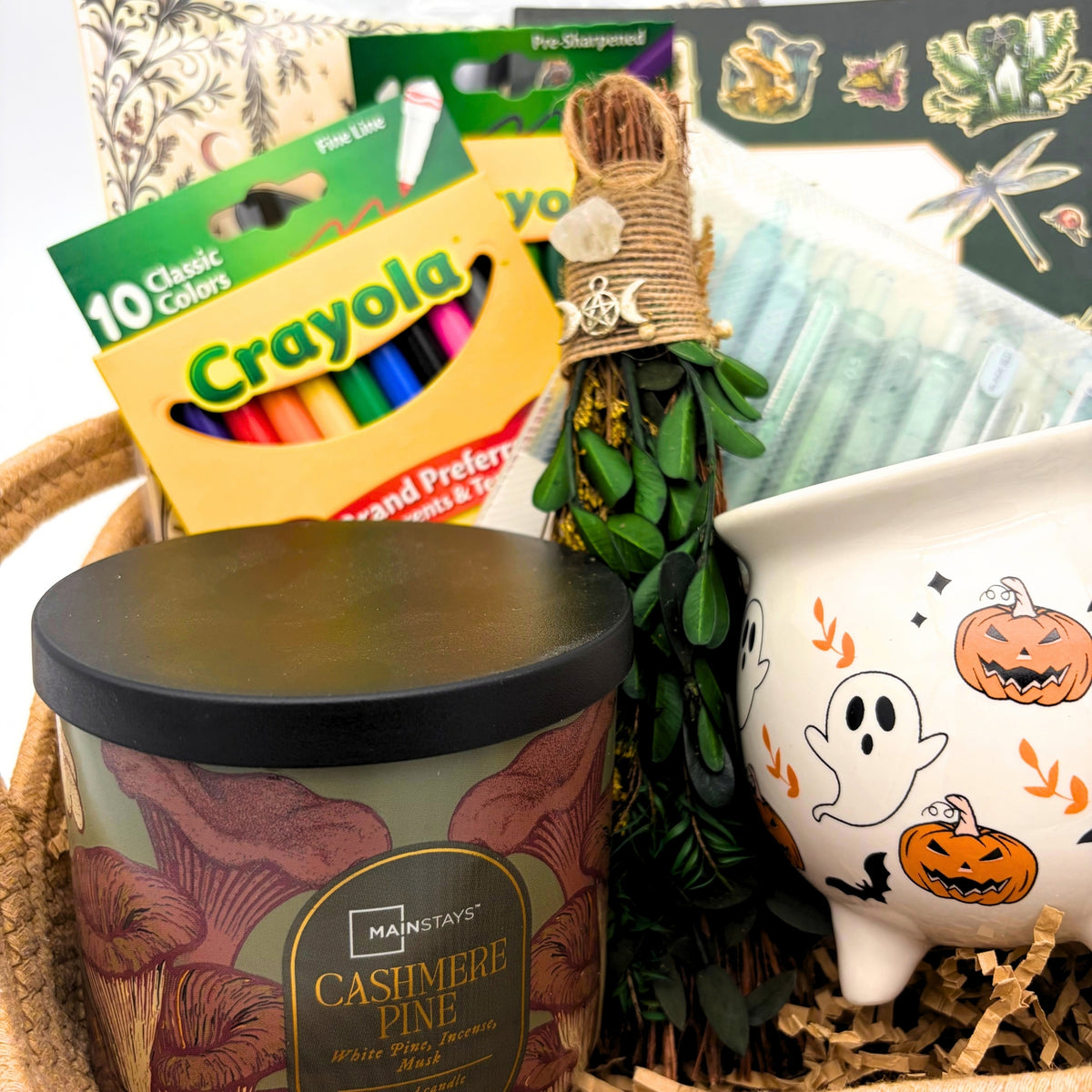 Close-up of the gift basket with a ghost and pumpkin cauldron mug, mini witch's broom with crystal and moon charm, and fine line markers