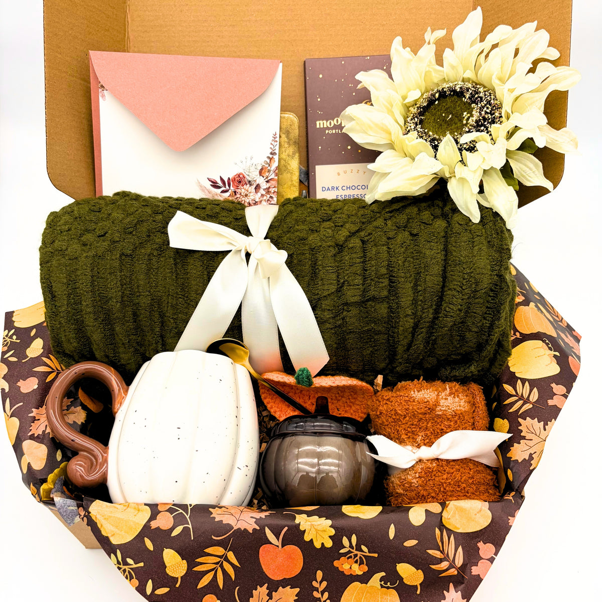 Close view of the olive green cozy fall gift box showing the knit throw blanket, pumpkin mug with gold spoon and crocheted pumpkin coaster