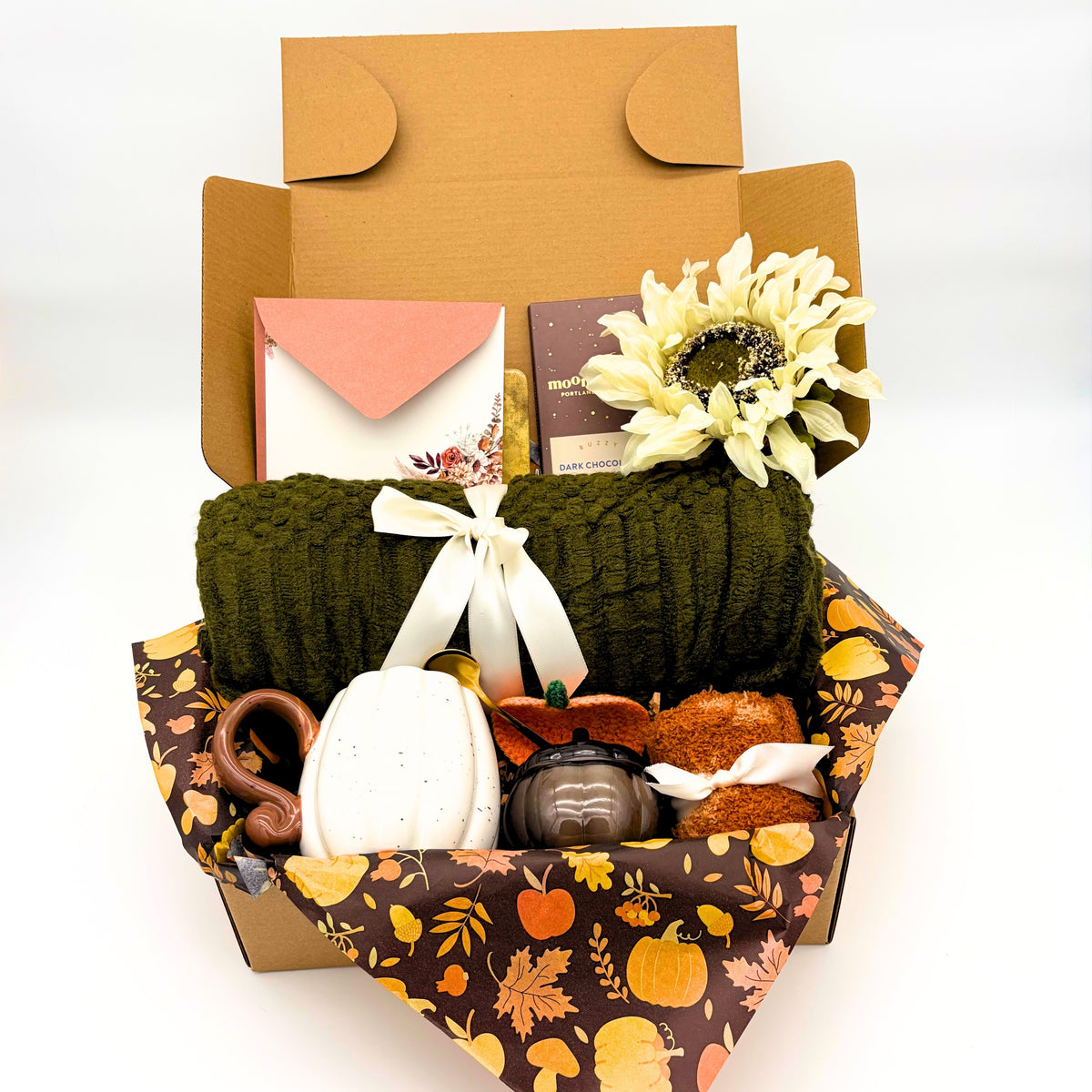 Cozy fall gift box with an olive green knit throw blanket, cream sunflower, white pumpkin mug, candle and fuzzy socks packed in a kraft gift box
