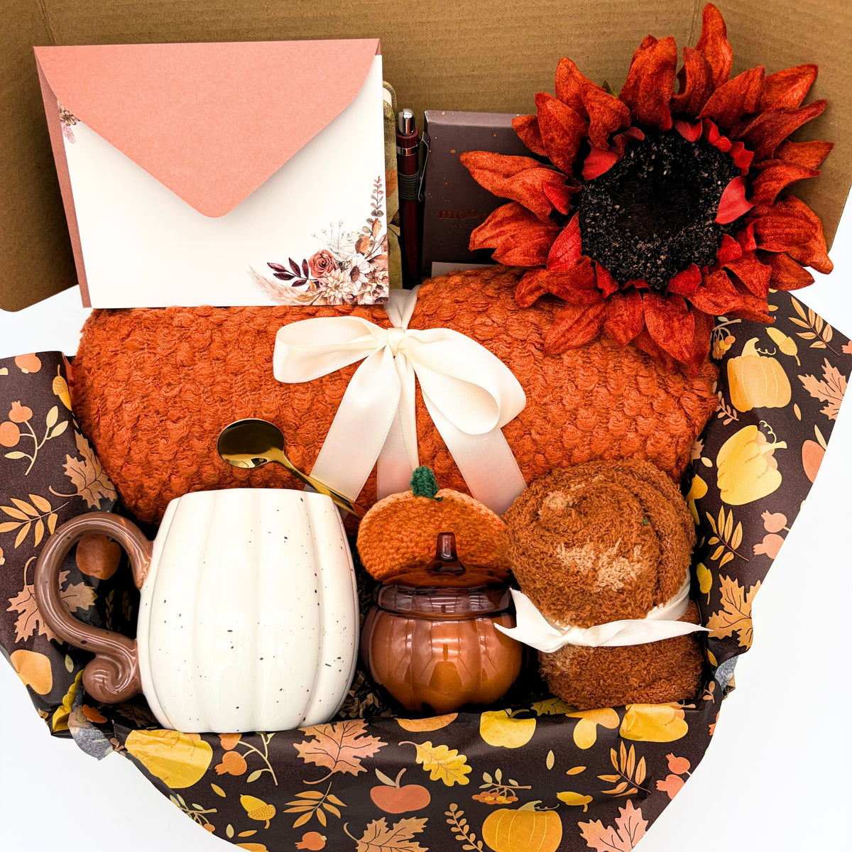 Close view of the orange cozy fall gift box showing the knit throw blanket, white ceramic pumpkin, gold stirring spoon and crocheted pumpkin coaster