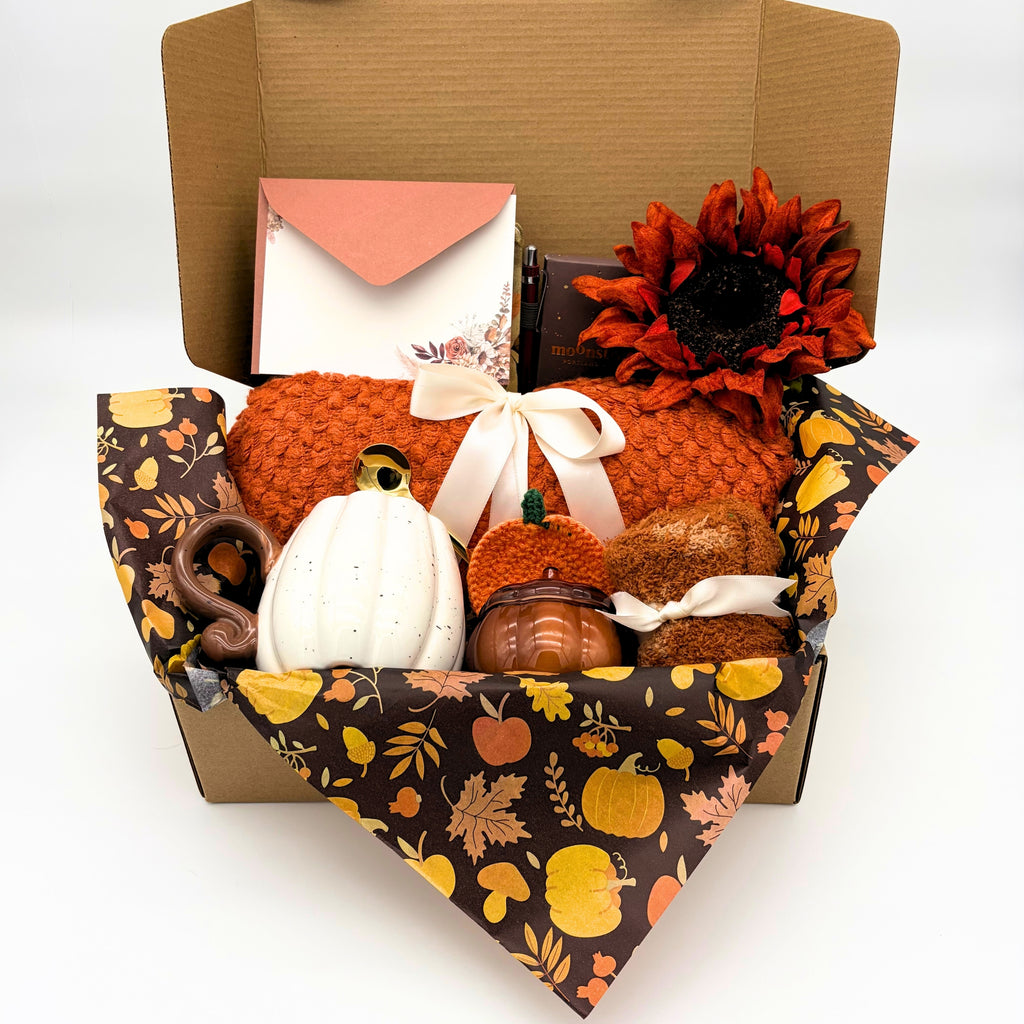 Cozy fall gift box with an orange knit throw blanket tied in a cream bow, white ceramic pumpkin mug, amber pumpkin candle, crocheted pumpkin coaster and fuzzy socks in a kraft box.