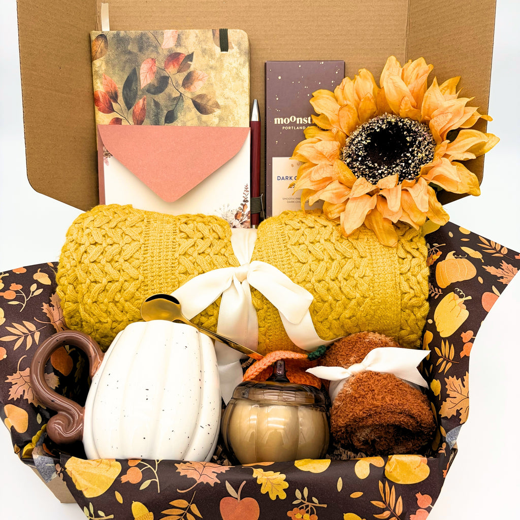 Close view of the yellow cozy fall gift box showing the knit throw blanket, ceramic pumpkin, pumpkin jar candle and fuzzy fall socks