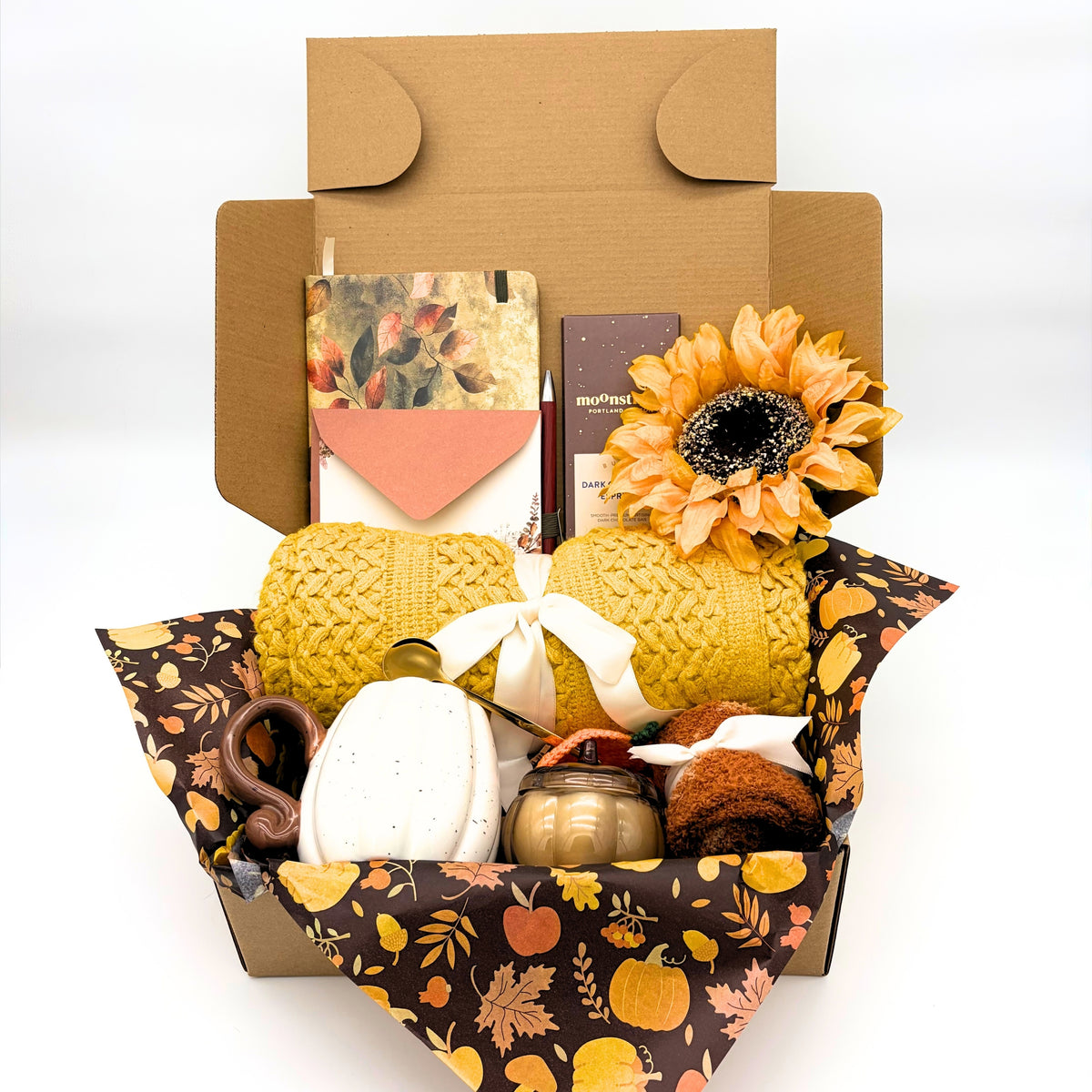 Cozy fall gift box with a yellow knit throw blanket, golden sunflower, pumpkin ceramic mug, dark chocolate bar and autumn notebook arranged in patterned tissue