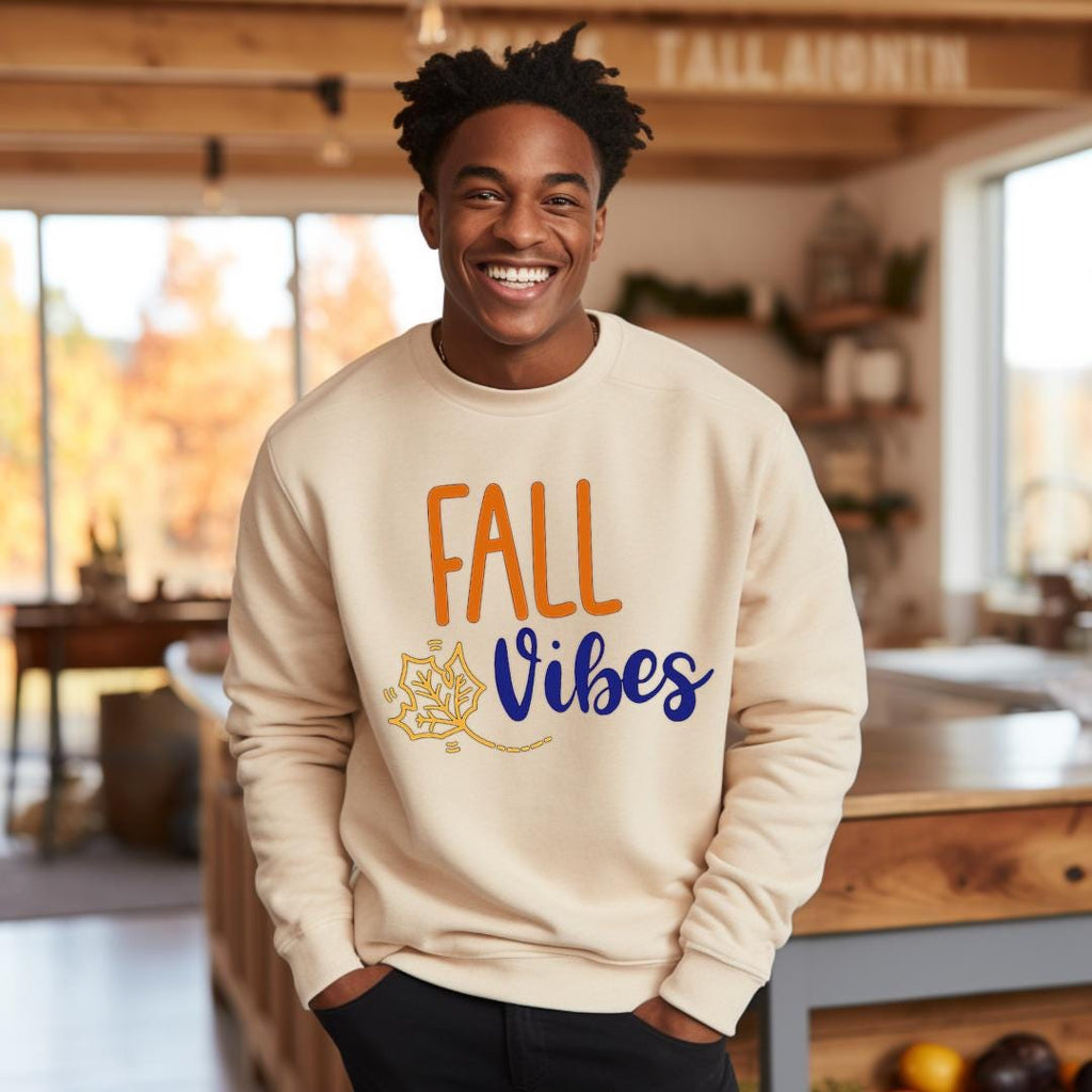"Fall Vibes" with "Fall" in orange block letters and "Vibes" in navy script above a decorative orange and navy leaf on a beige sweatshirt