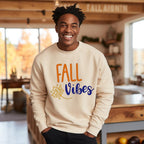 "Fall Vibes" with "Fall" in orange block letters and "Vibes" in navy script above a decorative orange and navy leaf on a beige sweatshirt