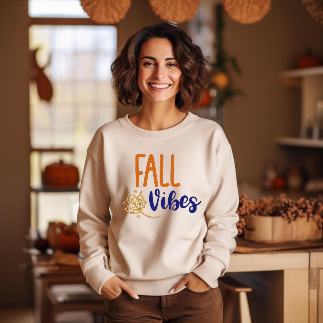 "Fall Vibes" with "Fall" in orange block letters and "Vibes" in navy script above a decorative orange and navy leaf on a beige sweatshirt