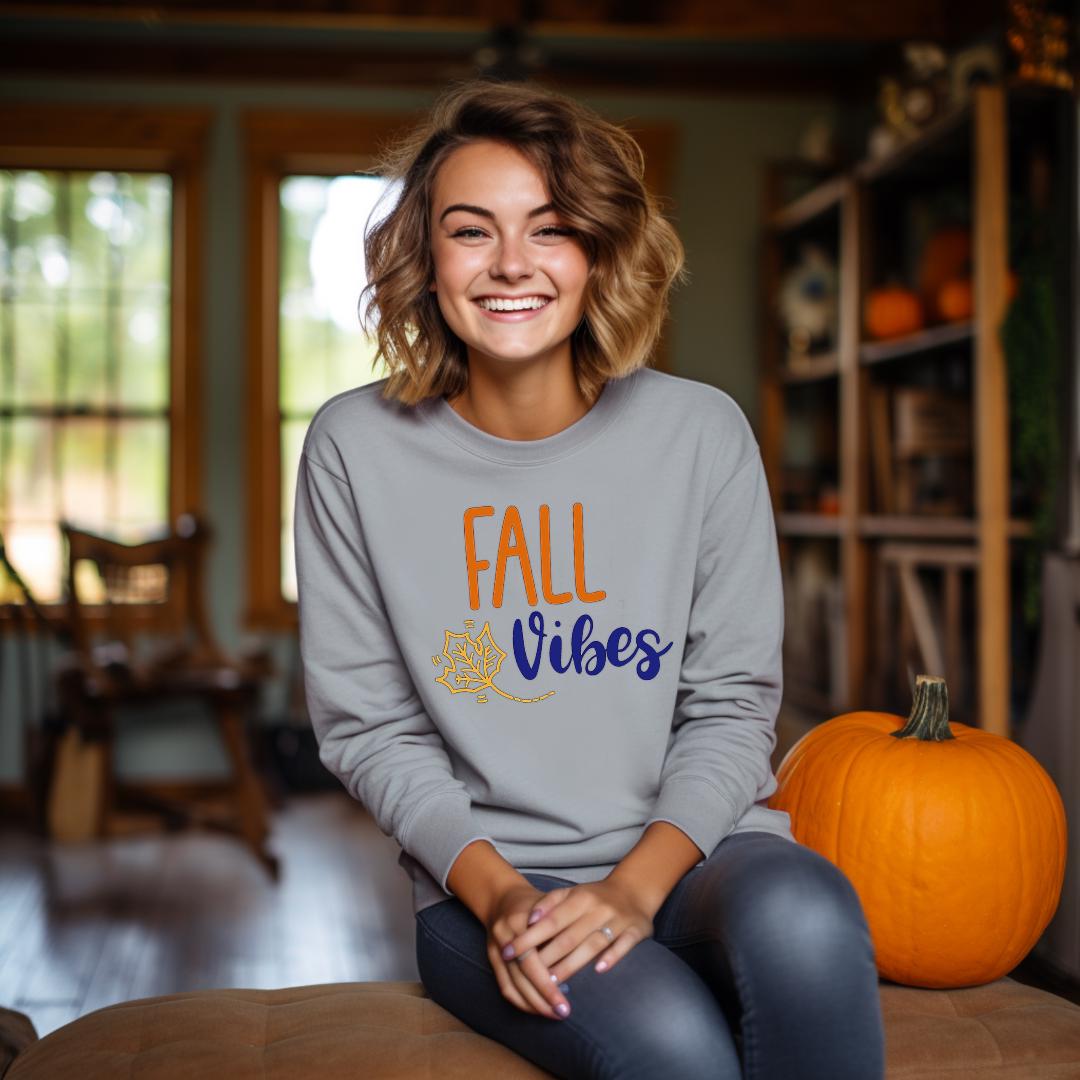 "Fall Vibes" with "Fall" in orange block letters and "Vibes" in navy script above a decorative orange and navy leaf on a gray sweatshirt