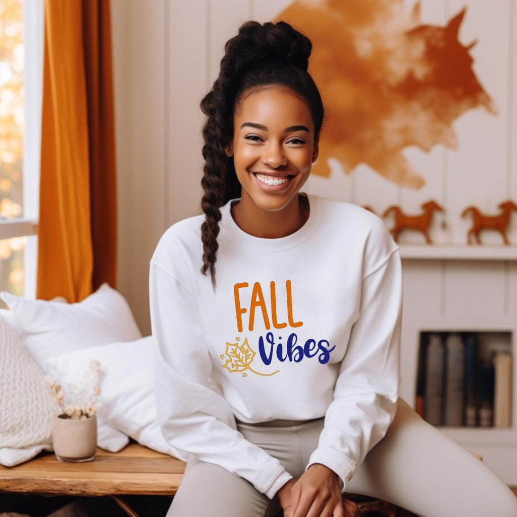 "Fall Vibes" with "Fall" in orange block letters and "Vibes" in navy script above a decorative orange and navy leaf on a white sweatshirt