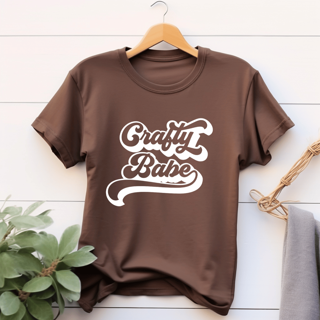Brown t-shirt with 'Crafty Babe' text on a wooden hanger against a white wooden background.